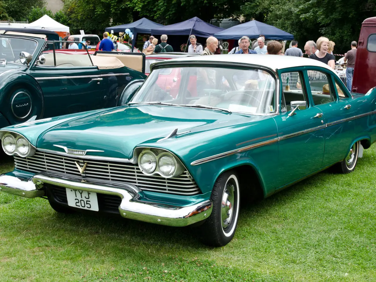 In this image there are vintage cars parked on the ground. There is grass on the ground. Behind the...