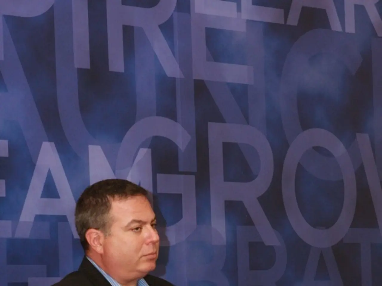 In the picture there is a man sitting on a chair and behind him there are a lot of alphabets on a...