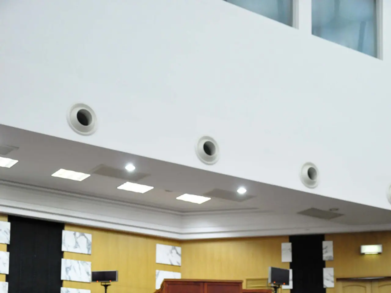 This is looking like a meeting hall. Many people are sitting on chairs. In front of them on table...