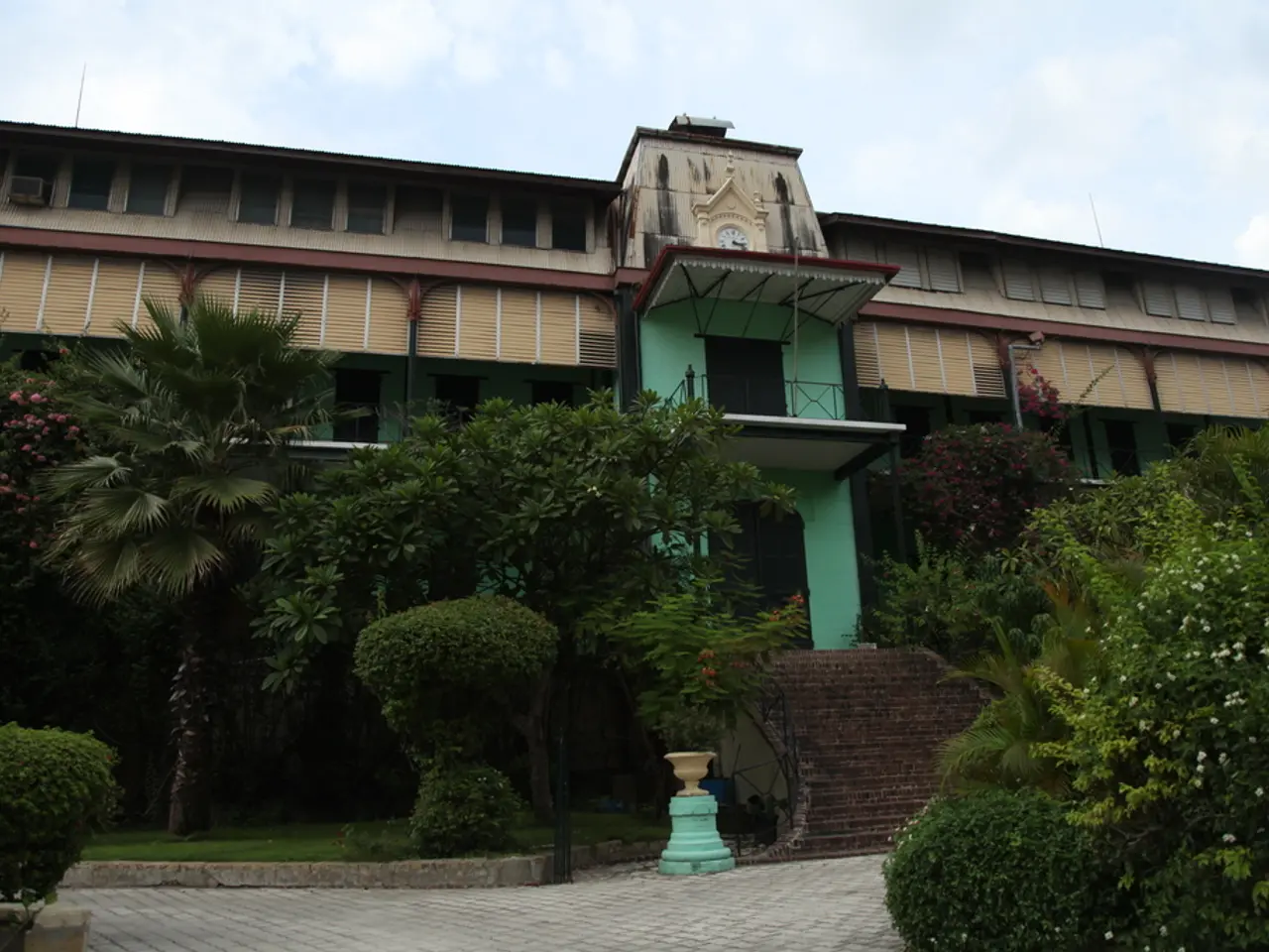 In this image there is a building, railings, steps, grass, plants, trees, pole, cloudy sky and...