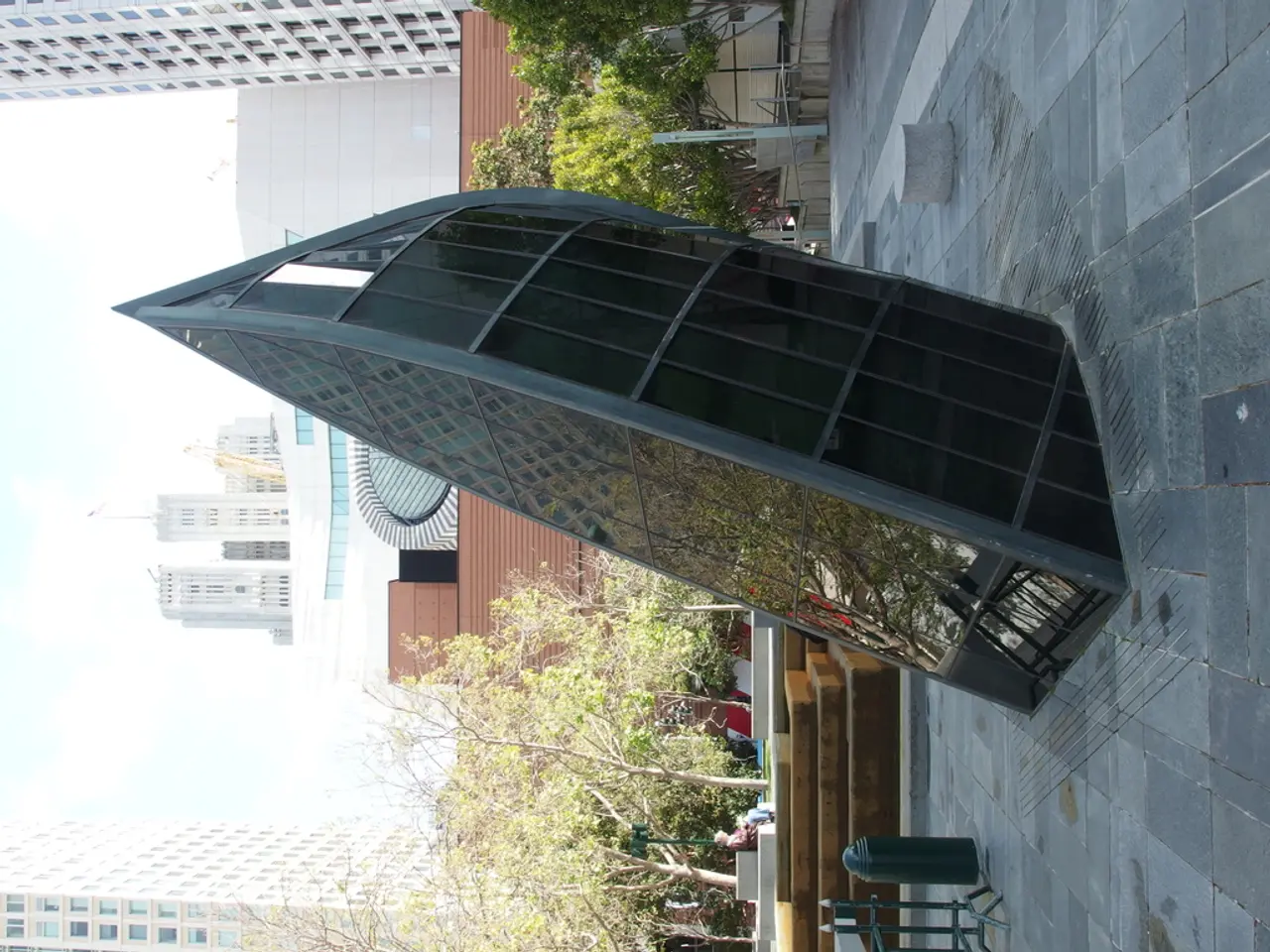 In this image we can see an architecture with glass. Behind it trees, poles and buildings are...