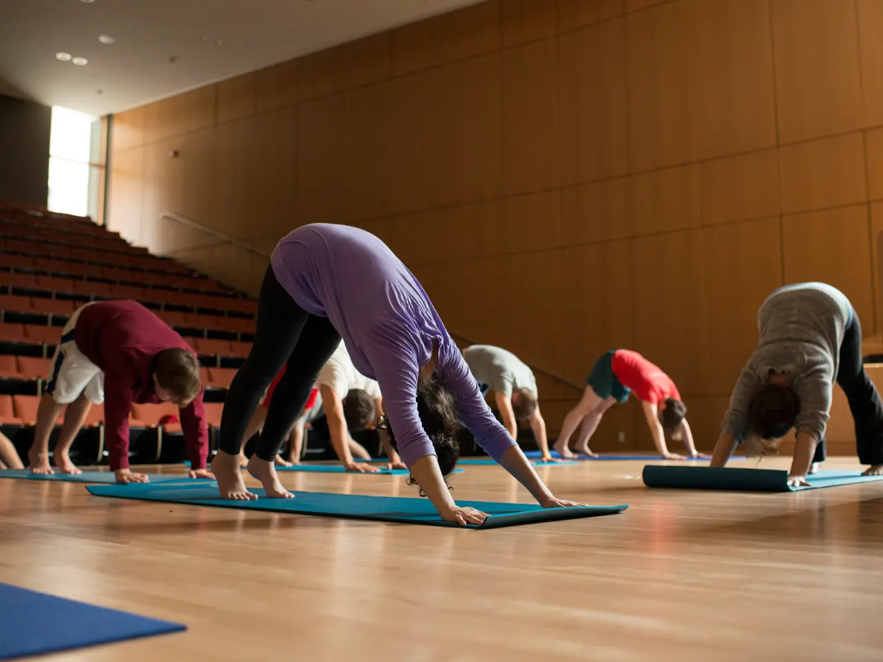 The image shows a group of people doing yoga in a large room with a wooden floor, a wall in the...