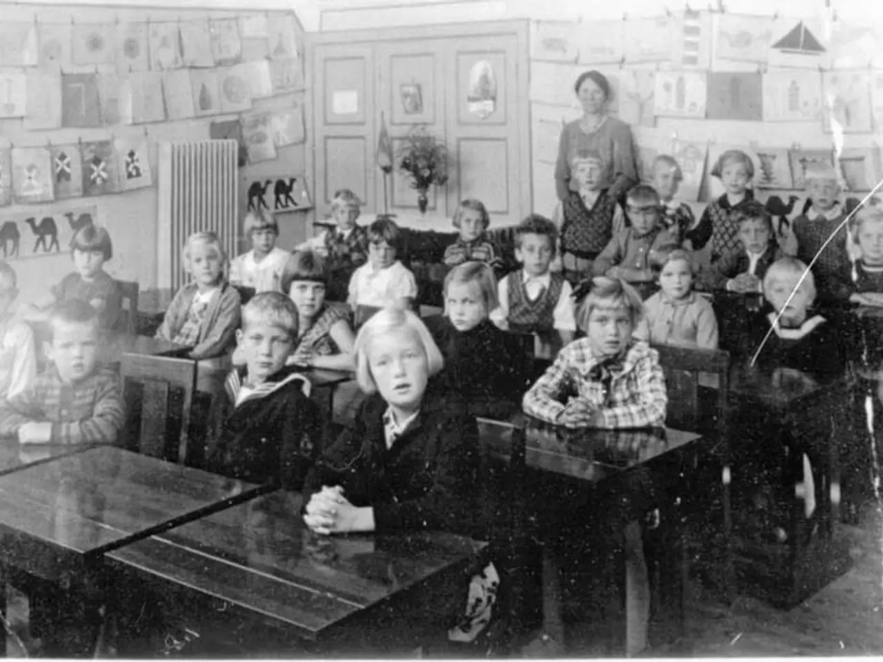 The image shows a black and white photo of a classroom full of children sitting on benches and some...