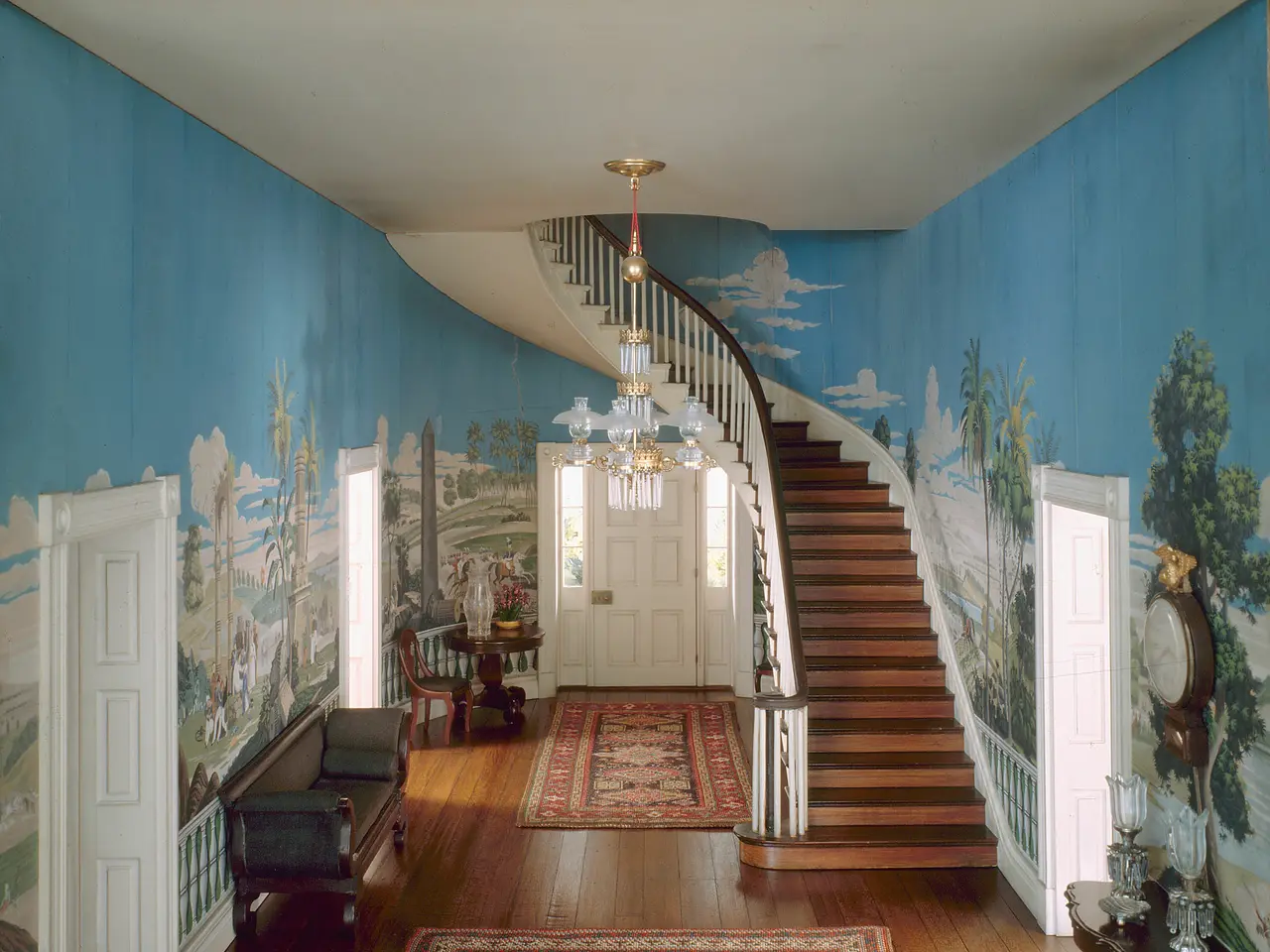 The image shows a foyer with blue walls and wood floors, a staircase with railings, a chandelier...