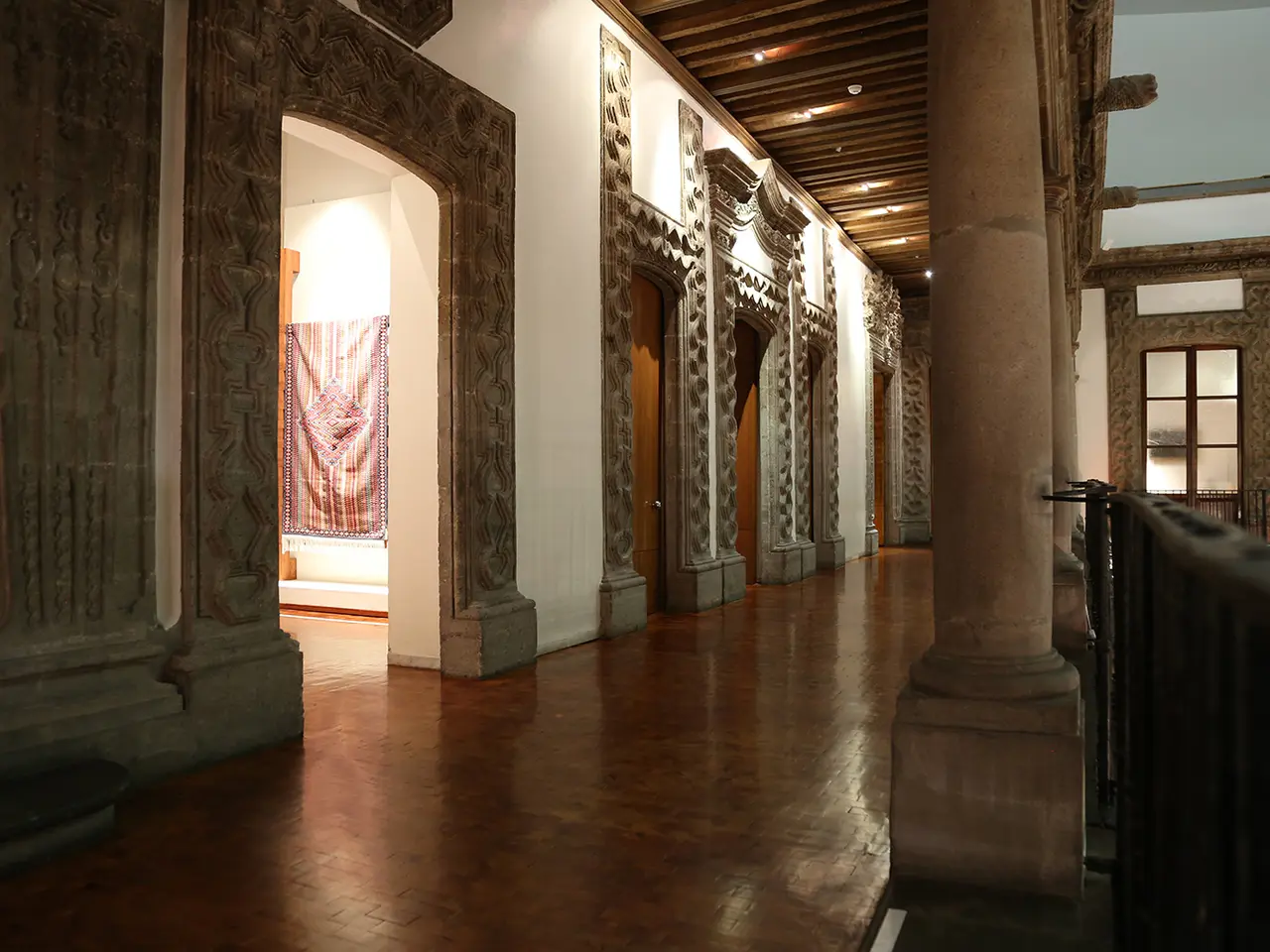 The image shows a large room with wooden floors, pillars, railings, walls, windows, lights, and a...