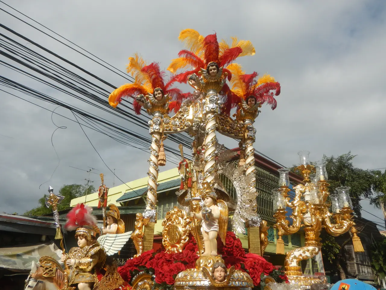 The image shows a float in a carnival parade with people on it, surrounded by buildings, trees,...