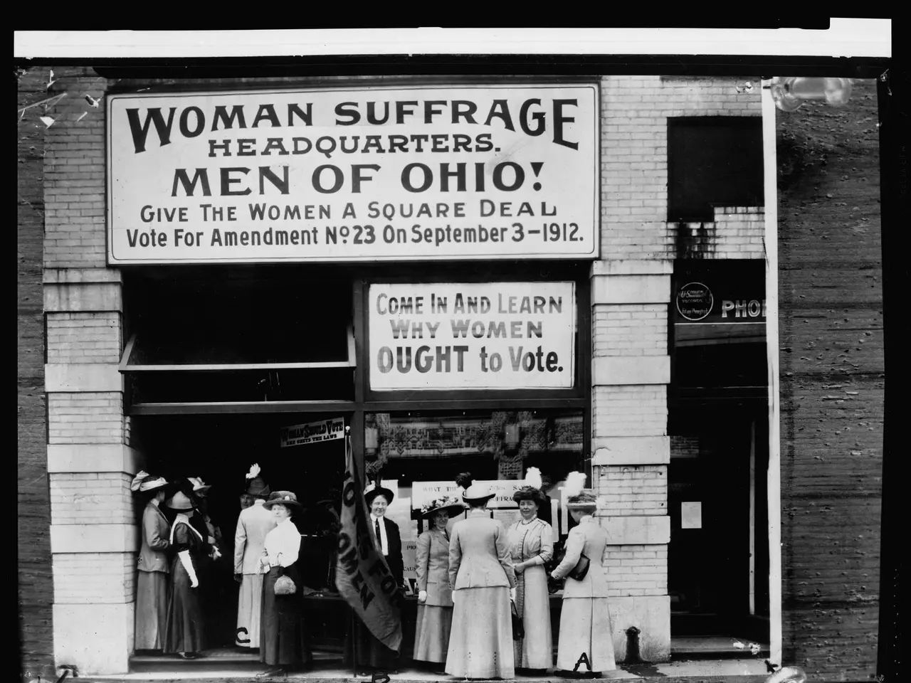 The image shows a group of women standing in front of a building with a sign that reads "Women...