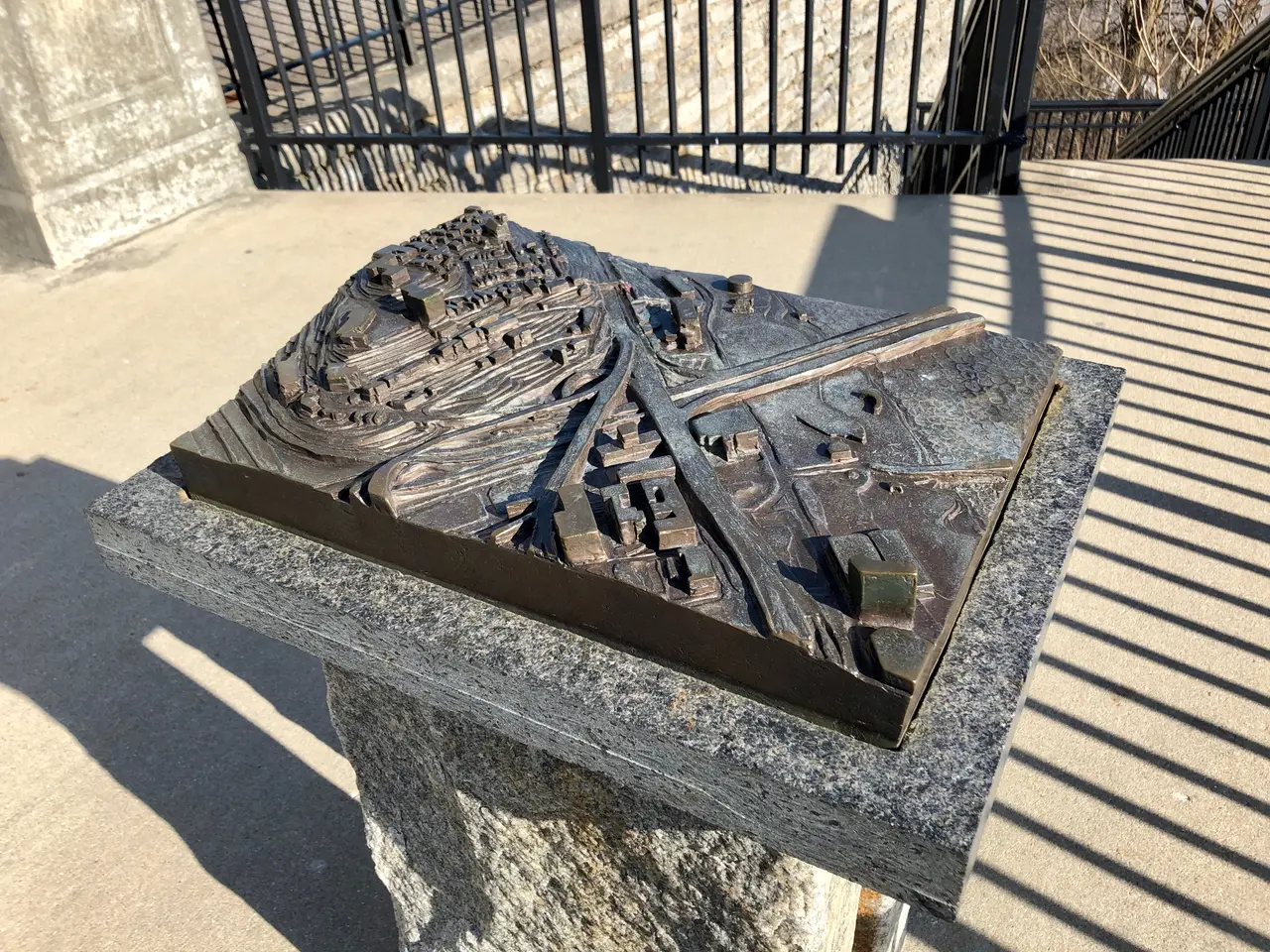 The image shows a bronze sculpture of a city on top of a stone pedestal, with a railing in the...