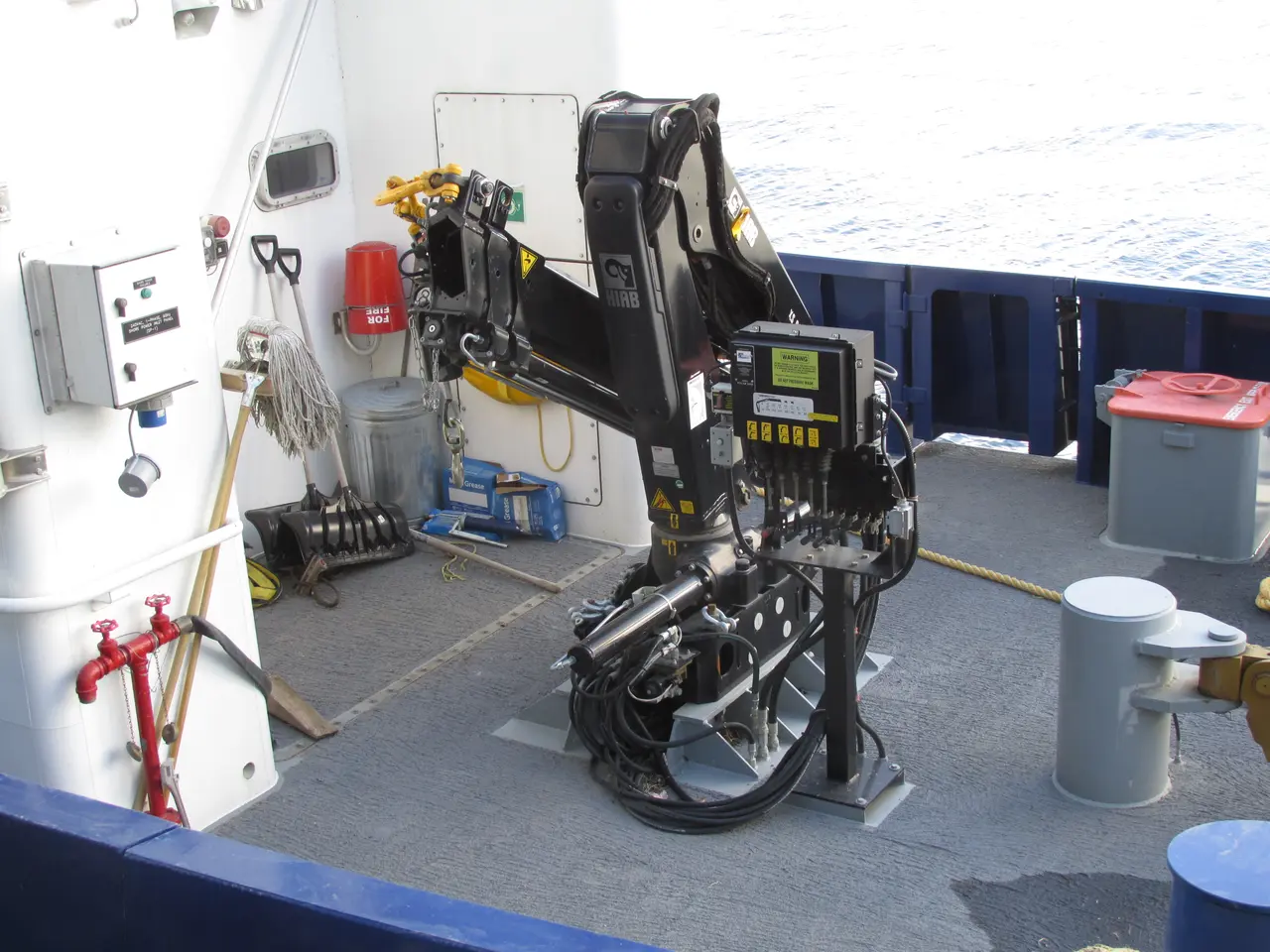 The image shows a robotic arm on the deck of a boat, surrounded by machines, wires, buckets, and...