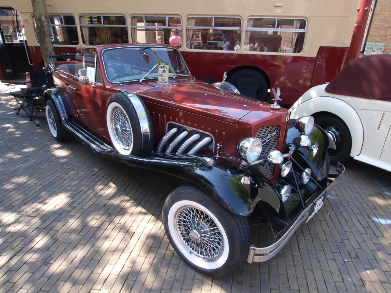 The image shows a vintage car parked next to a double decker bus on a cobblestone street. There are...