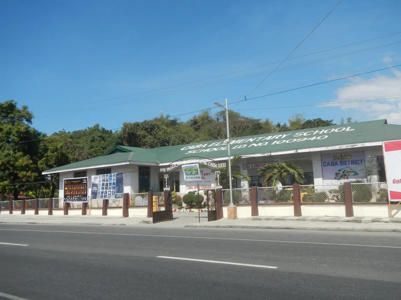 The image shows a school building with a green roof on the corner of a street, surrounded by a...