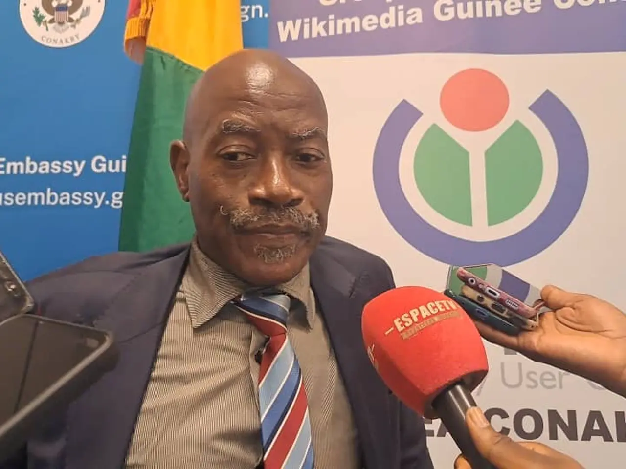 The image shows a man in a suit and tie talking to reporters, with a person's hand holding a...