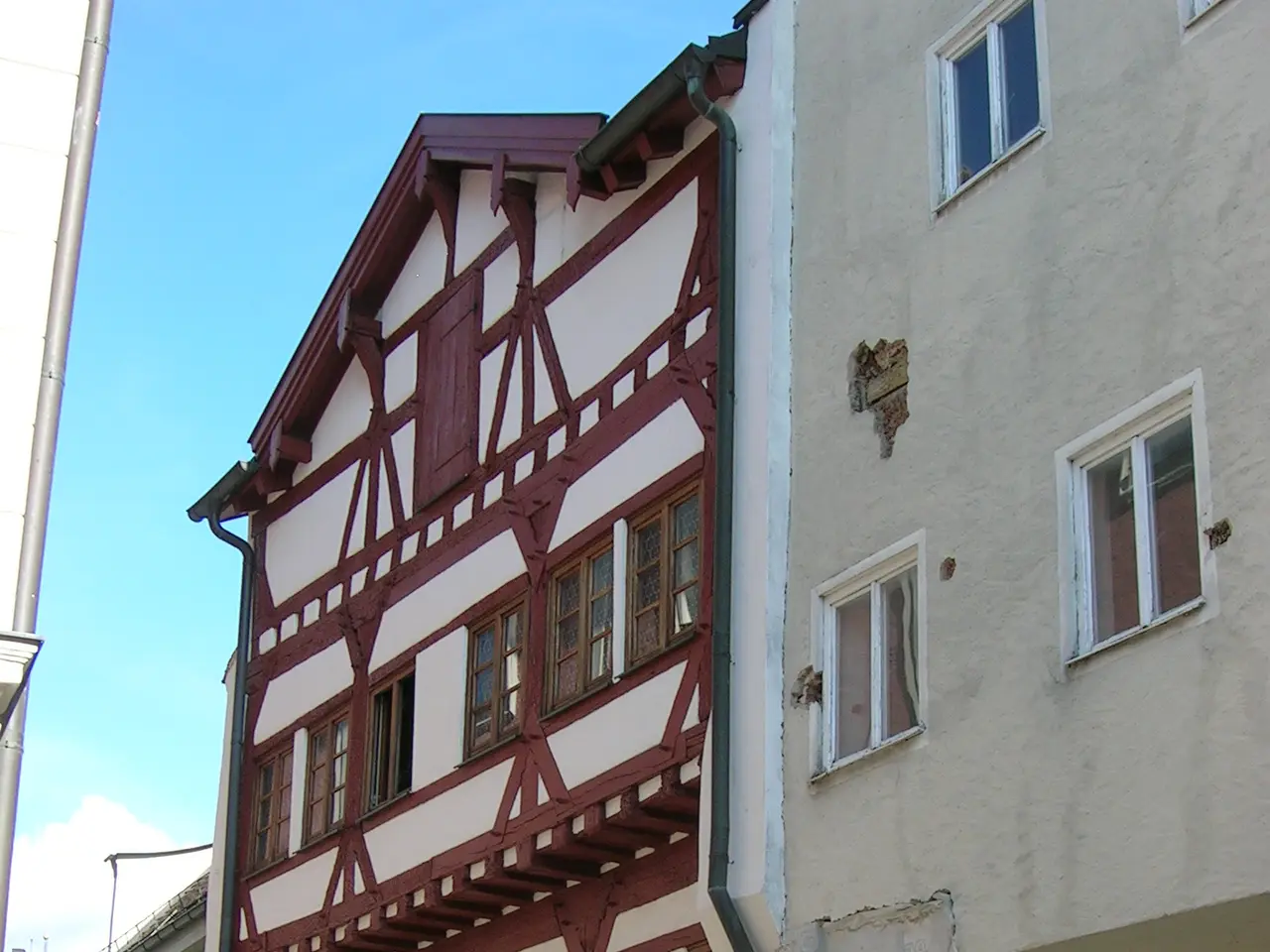The image shows a half-timbered building in the middle of a cobblestone street, surrounded by trees...