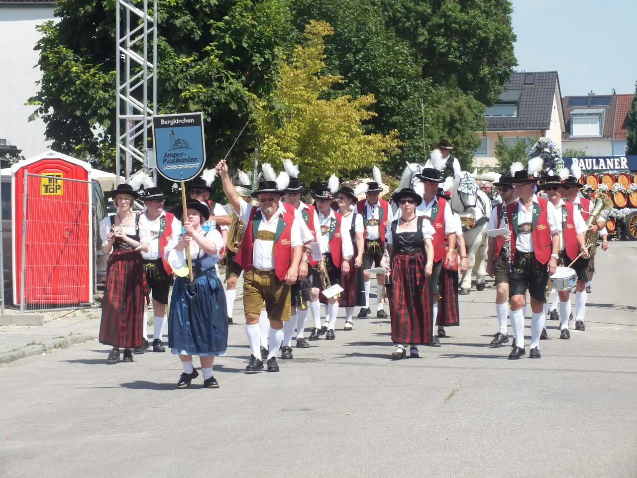 The image shows a group of people in traditional Bavarian clothing walking down a street, some of...