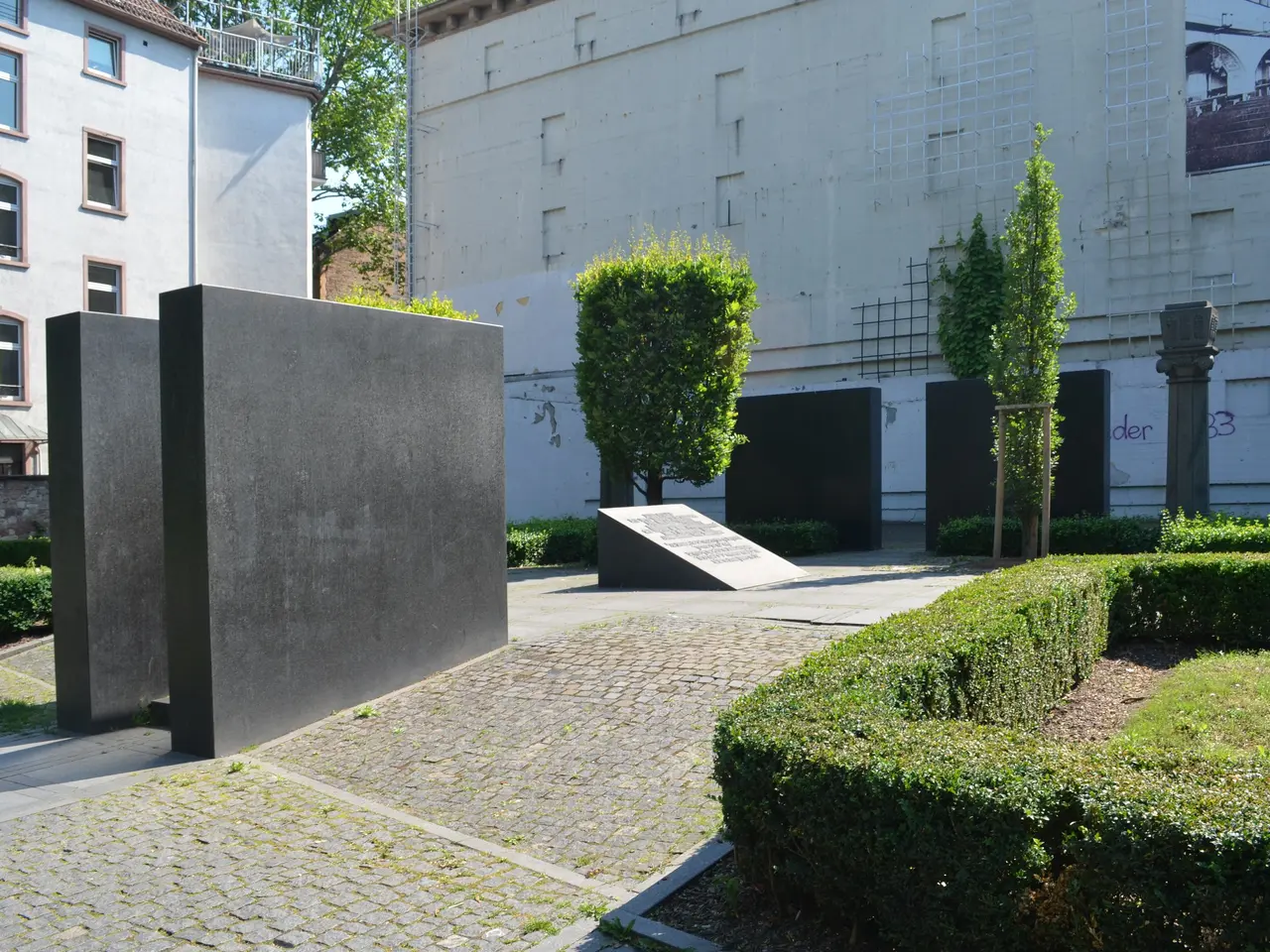 The image shows a garden with hedges and bushes in front of a building, surrounded by trees and a...