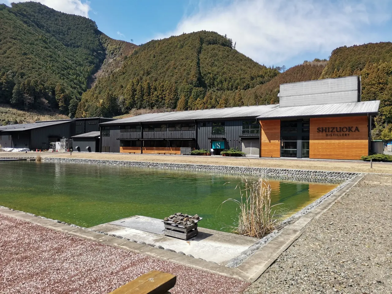 The image shows a large building with a pond in front of it, surrounded by lush green plants and...