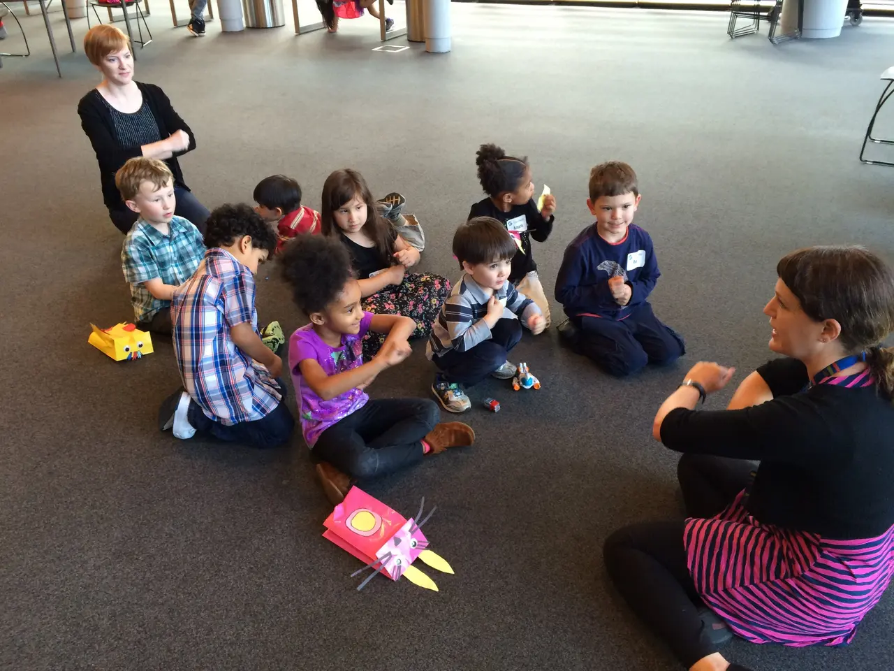 The image shows a group of children sitting on the floor in front of a teacher, surrounded by toys....