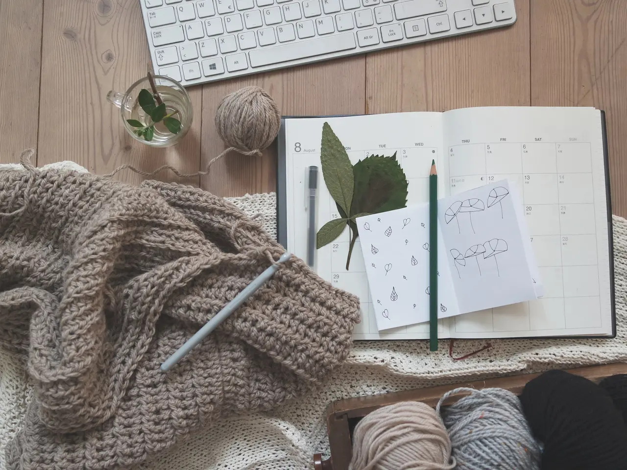 The image shows a wooden desk with a keyboard, a notebook, a pen, a cloth, a ball of yarn, a plant...