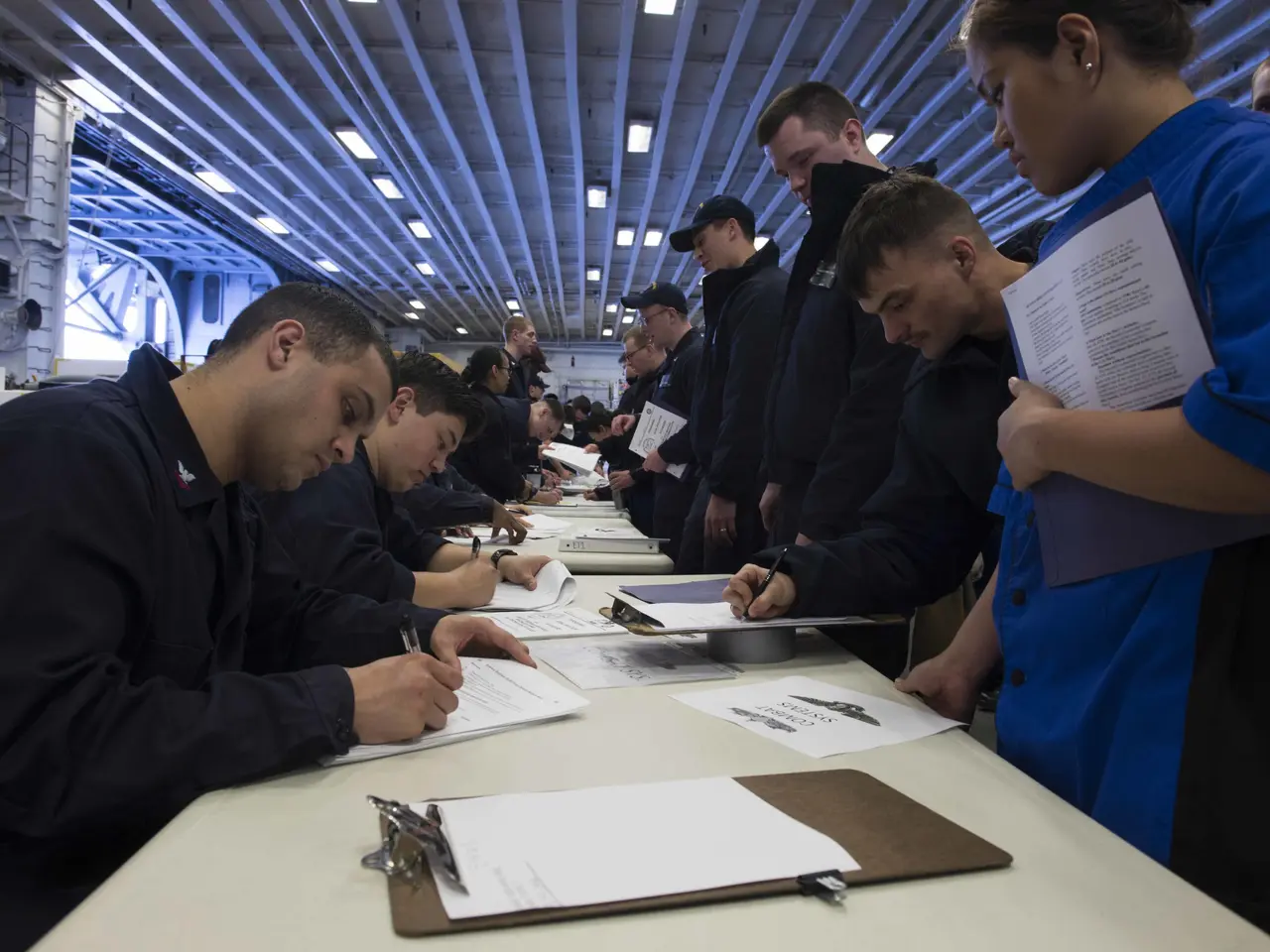 The image shows a group of people sitting at a table in a room, with papers and files spread out in...
