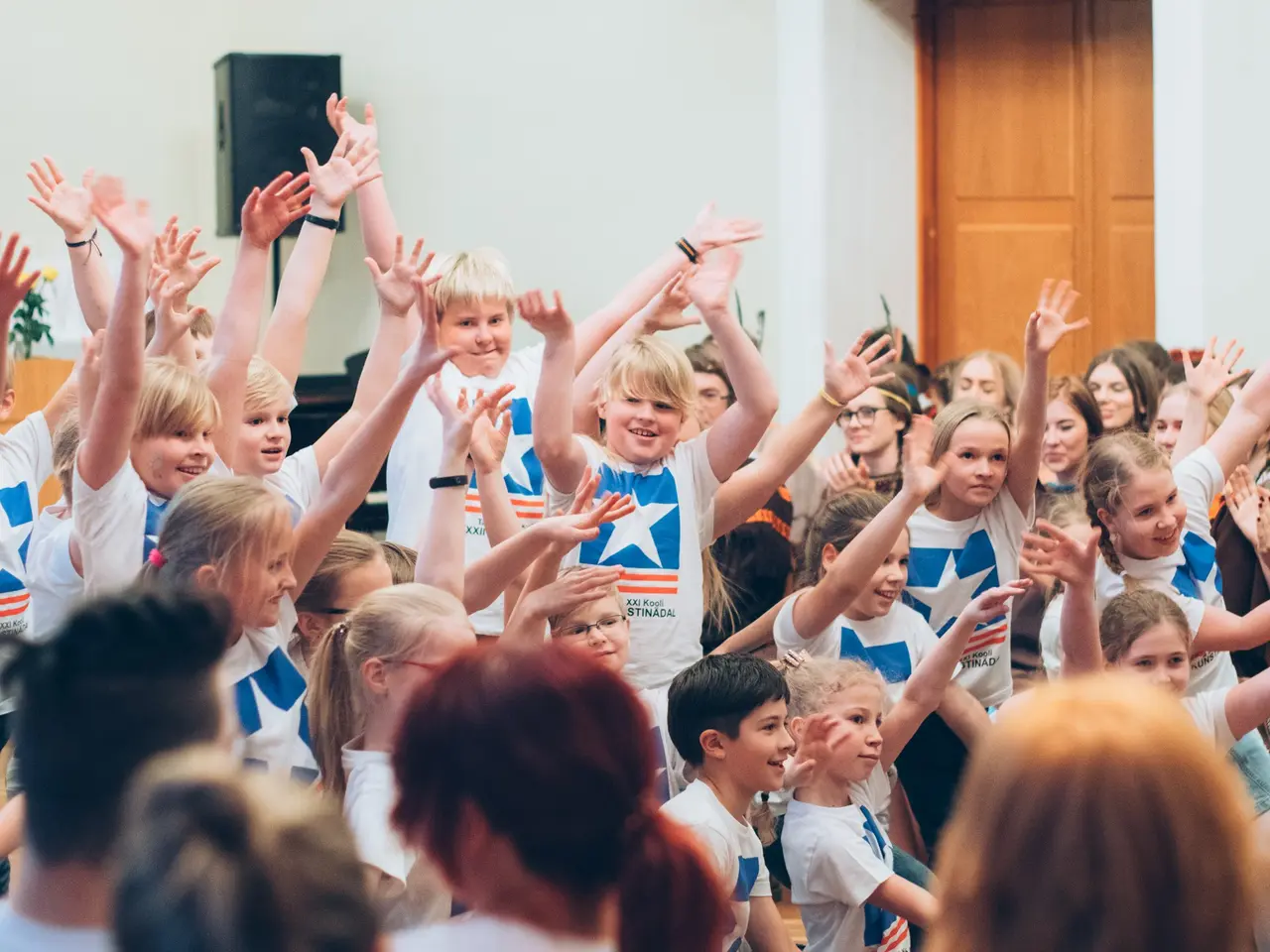 The image shows a group of children standing in front of a crowd with their hands in the air,...