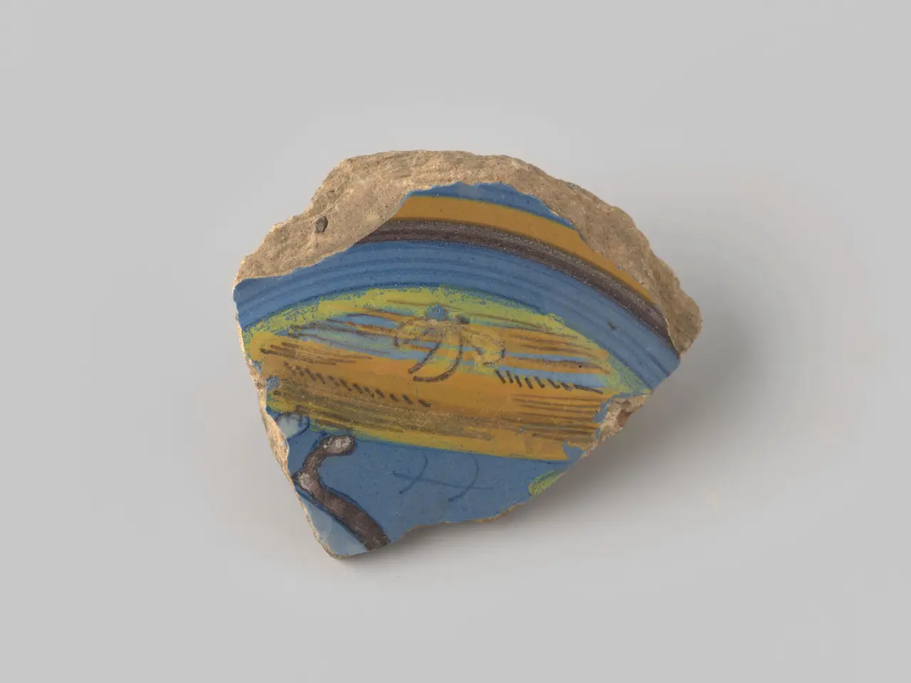 The image shows a small piece of pottery with a painting on it in blue, yellow, and brown colors...