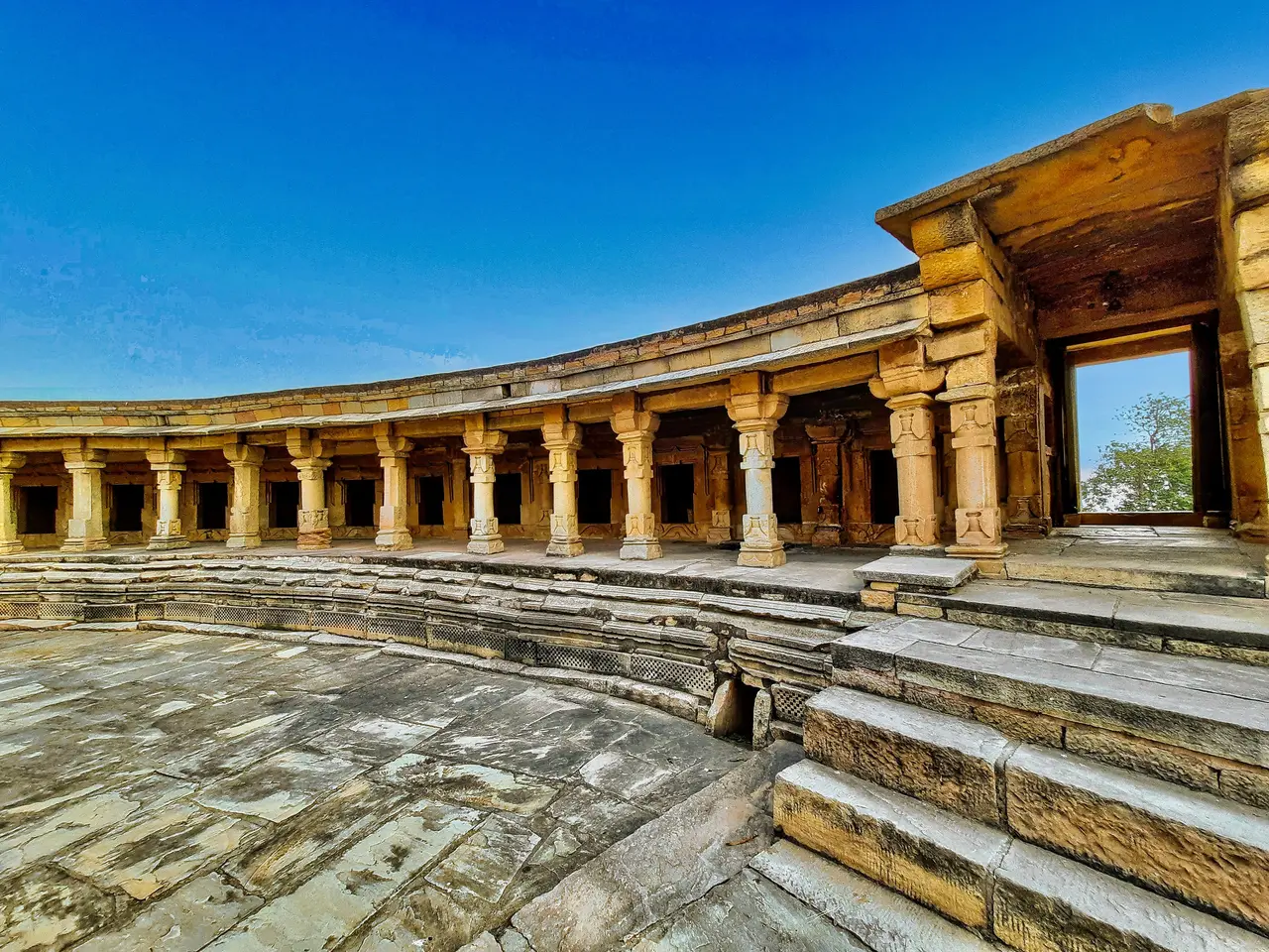 The image shows a large stone building with many pillars and steps in the middle of it, surrounded...
