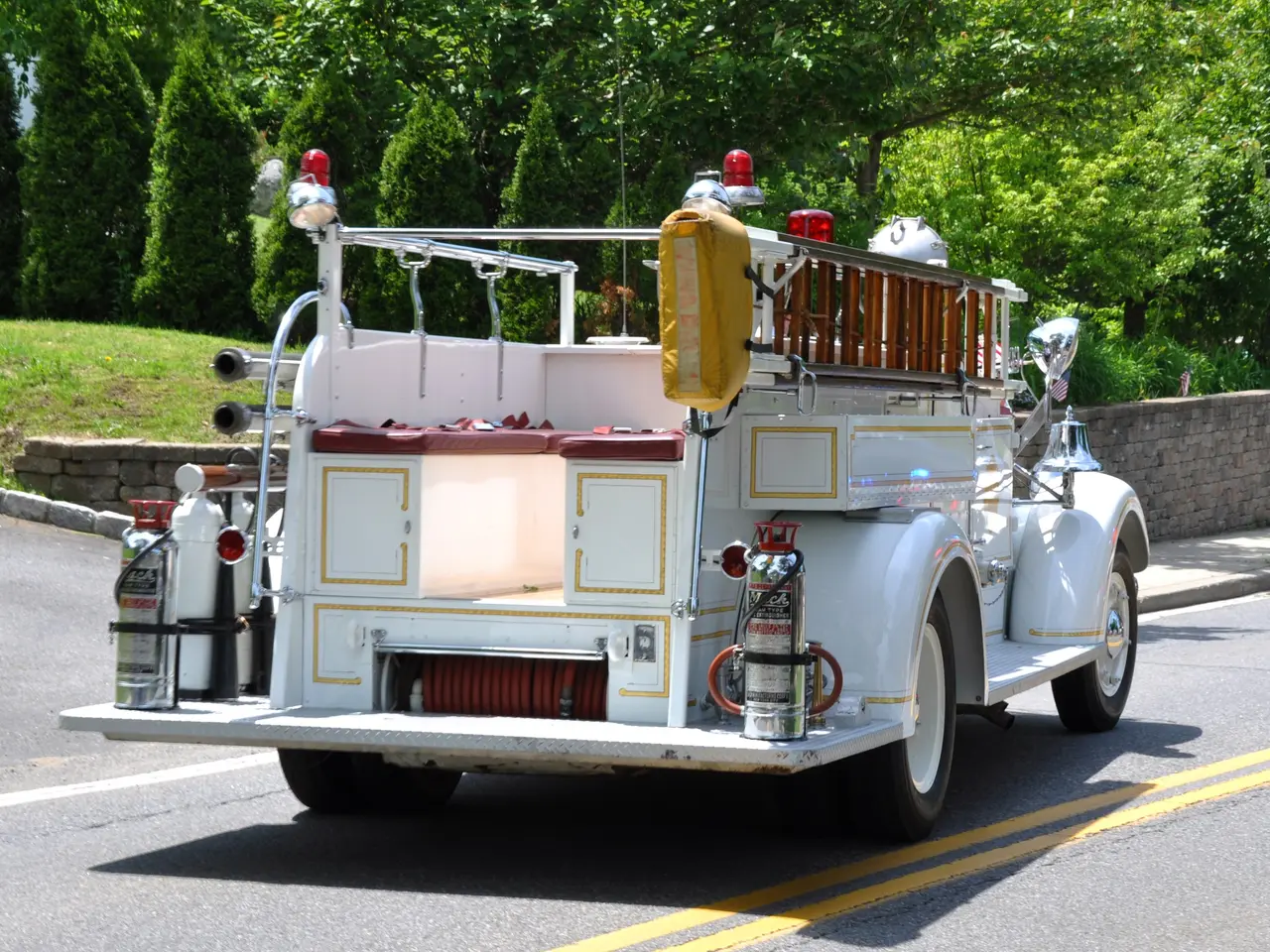 The image shows a white fire truck parked on the side of the road, surrounded by a wall, grass,...