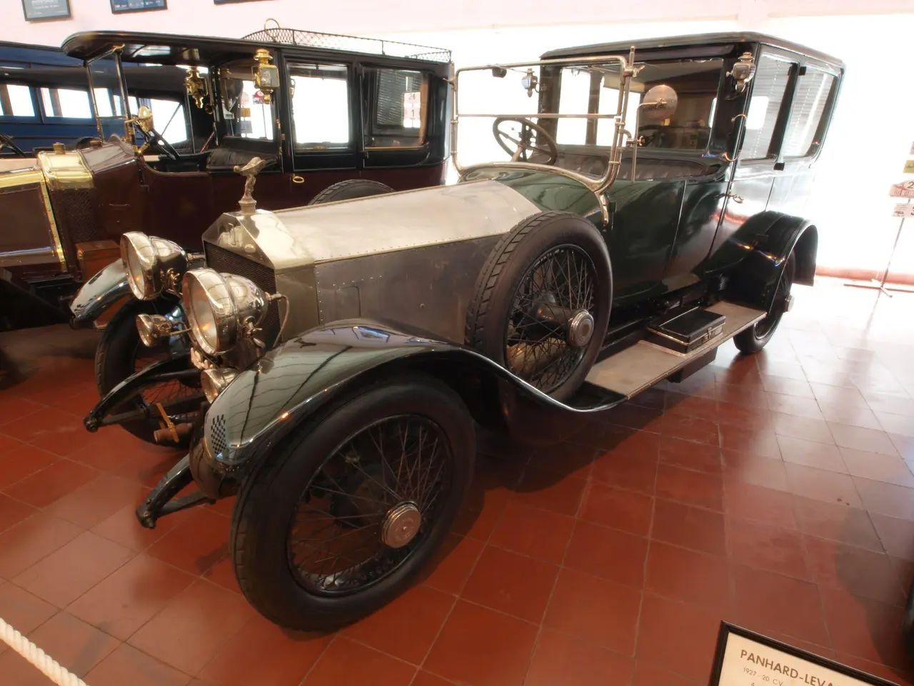 The image shows a vintage car on display in a museum, surrounded by boards with text, a rope, and...