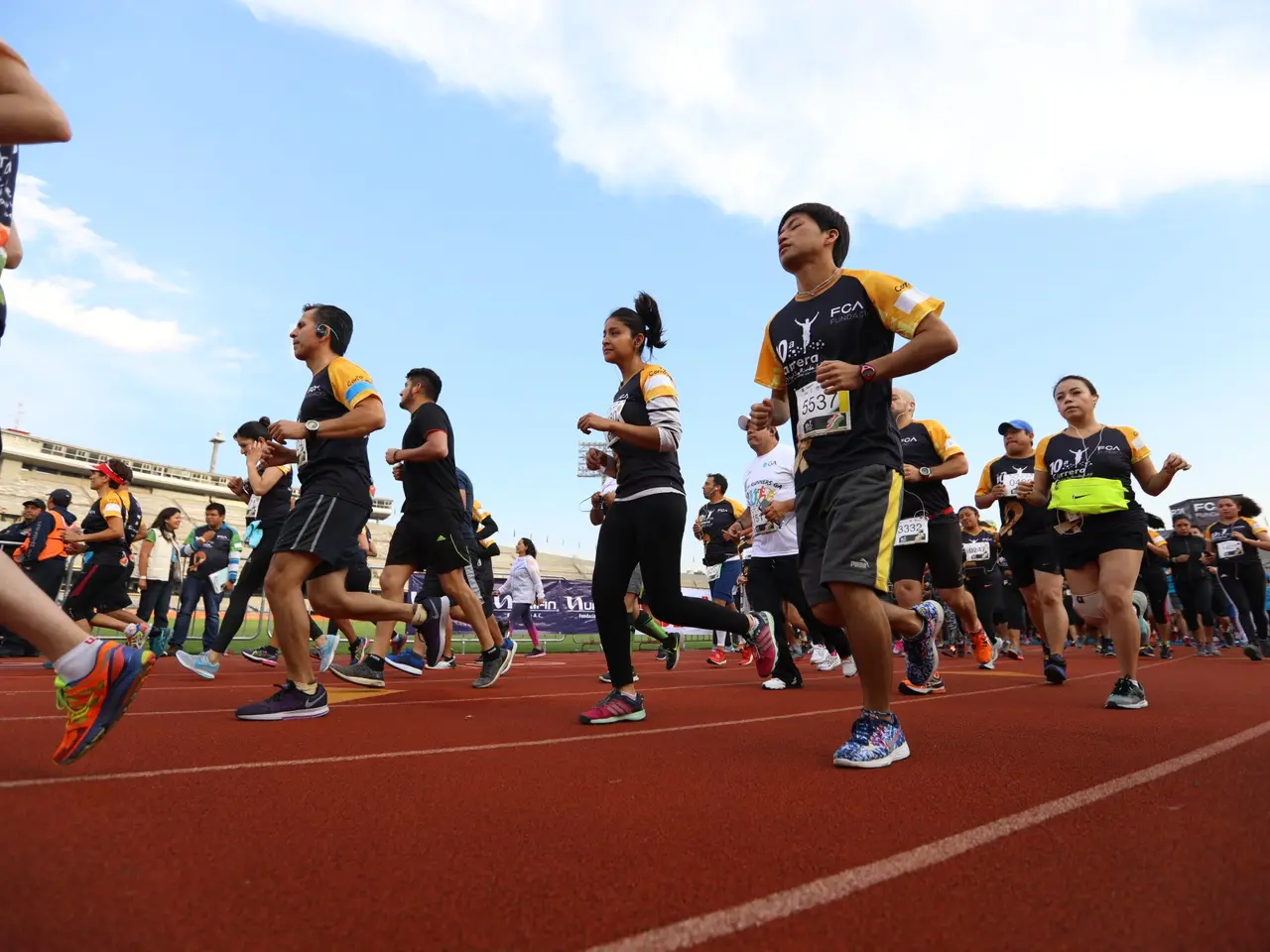 The image shows a group of people running on a track, with a crowd of people standing on the right...