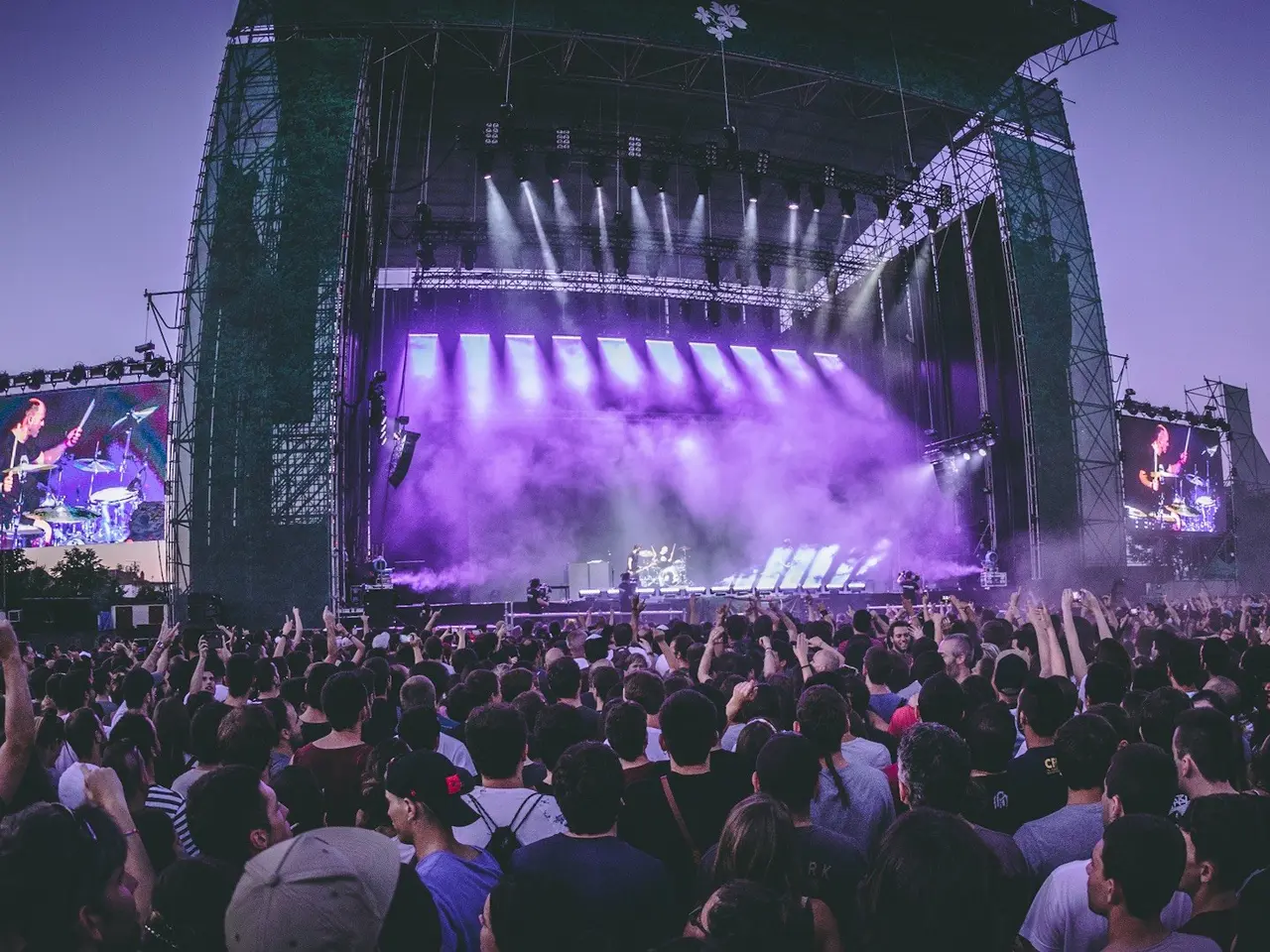 The image shows a large crowd of people gathered at a music festival, with a stage in the center of...