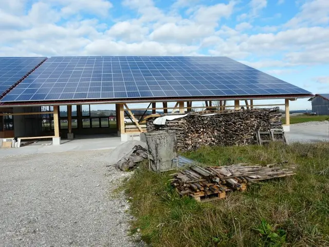 The image shows a building with solar panels on its roof, surrounded by grass and wooden logs. In...