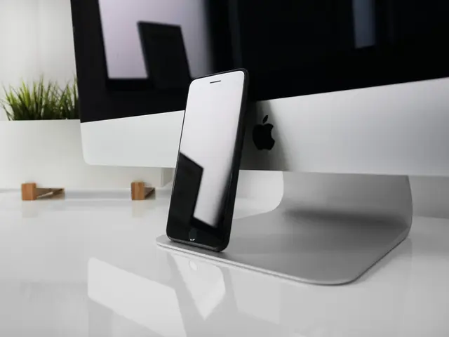 The image shows an iPhone sitting on top of a desk next to a computer monitor, with a houseplant in...