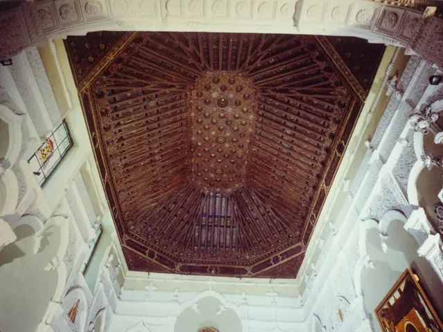 The image shows the interior of a building with a wooden ceiling, adorned with intricate carvings...