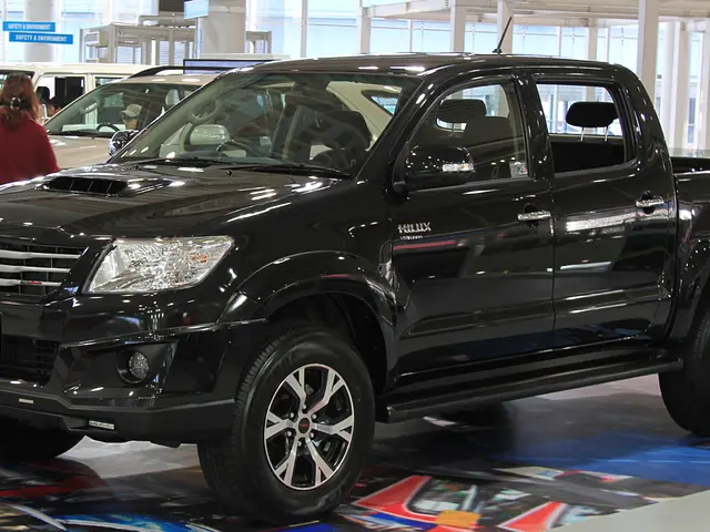 The image shows a black Toyota Hilux pickup truck on display at an auto show, surrounded by a group...