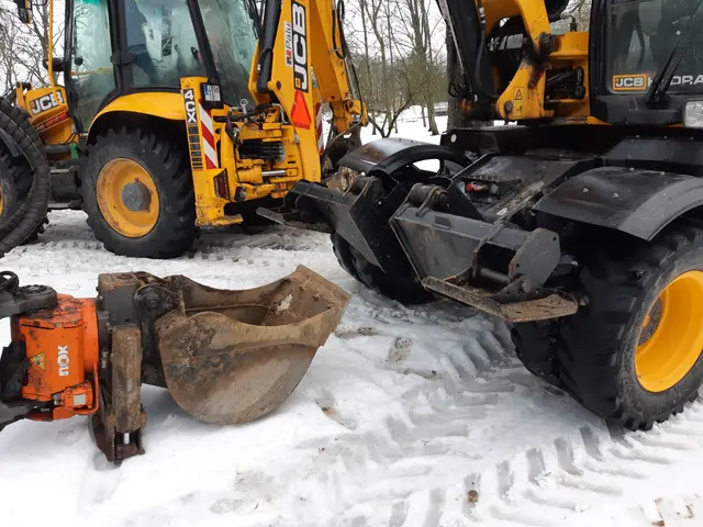 The image shows two JCB backhoes parked next to each other in the snow, surrounded by trees and a...