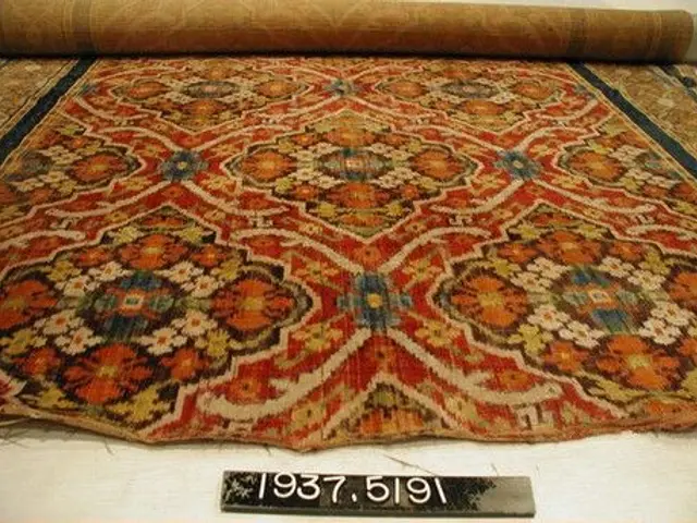 The image shows an antique Persian rug on display in a museum. The rug is laid out on the floor,...