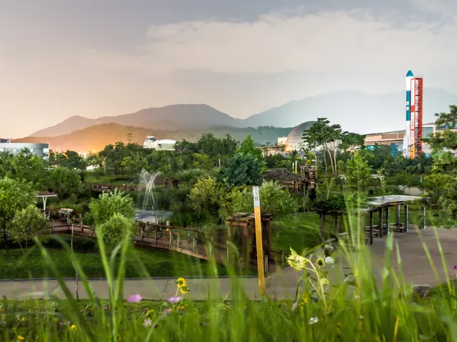 The image shows a park with lush green grass, vibrant flowers, tall trees, a bridge with railings,...