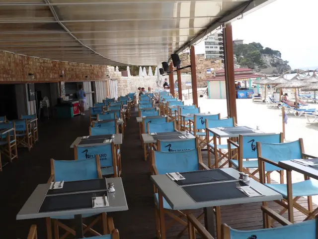 The image shows a restaurant with tables and chairs on the beach, surrounded by umbrellas, people,...