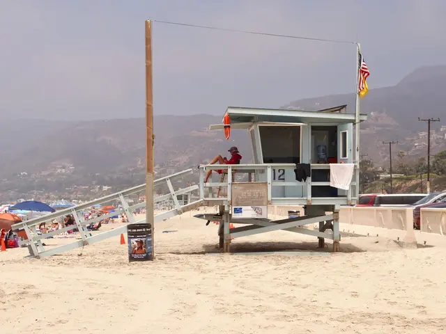 The image shows a lifeguard tower on the beach with people sitting on it, surrounded by sand,...