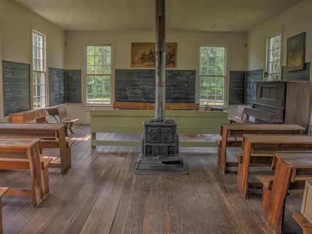The image shows an empty classroom with wooden desks and chairs arranged in neat rows, a pole in...