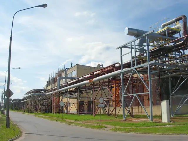 The image shows a large factory with a lot of pipes on the side of it, surrounded by buildings,...