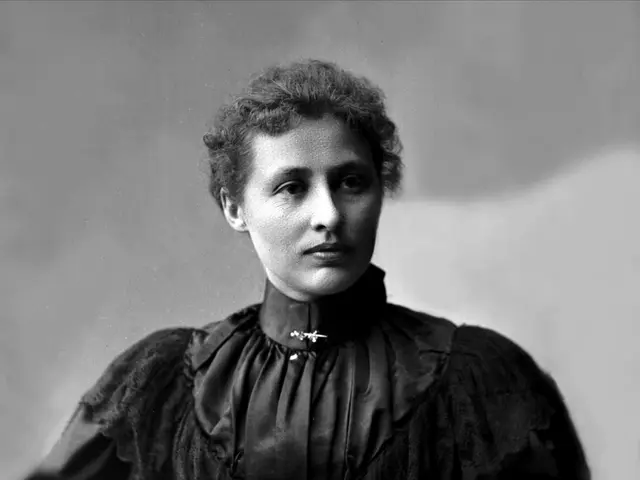 The image shows a black and white photo of a woman in a black dress, believed to be the first woman...