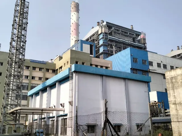 The image shows a large factory with a lot of buildings in the background, surrounded by a mesh...