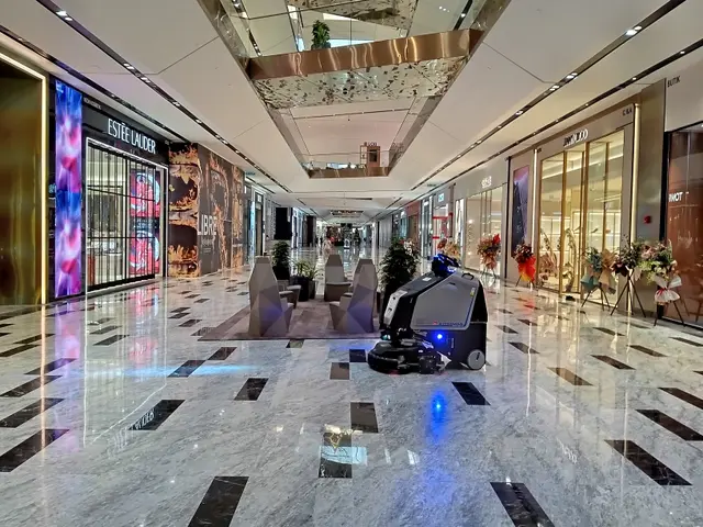 The image shows a robot cleaning the floor of a shopping mall, surrounded by houseplants, chairs,...