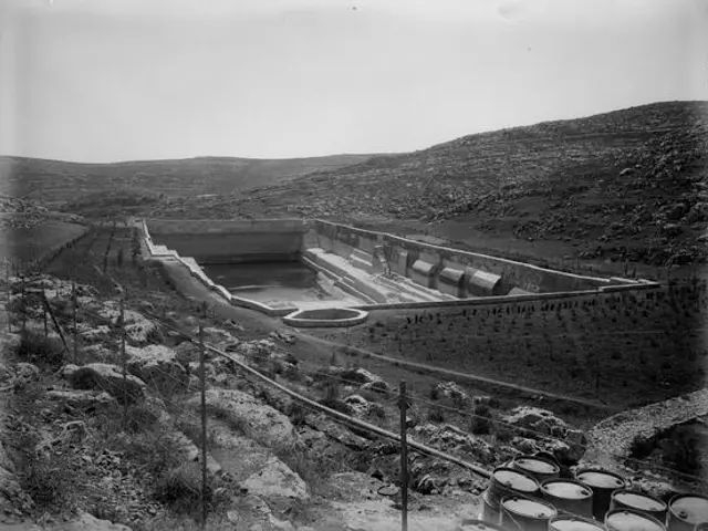 The image shows a black and white photo of a large dam in the middle of a field, surrounded by...