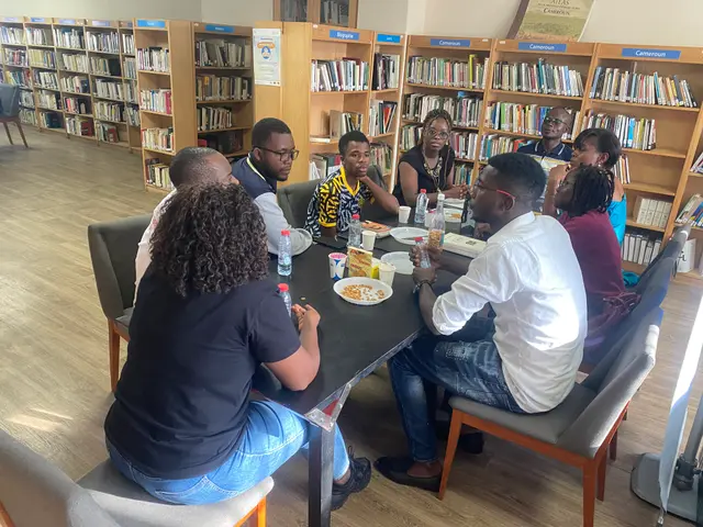 The image shows a group of people sitting around a table in a library, surrounded by bookshelves...