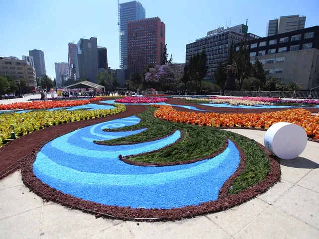 The image shows a vibrant flower garden in the middle of a city square, surrounded by buildings,...