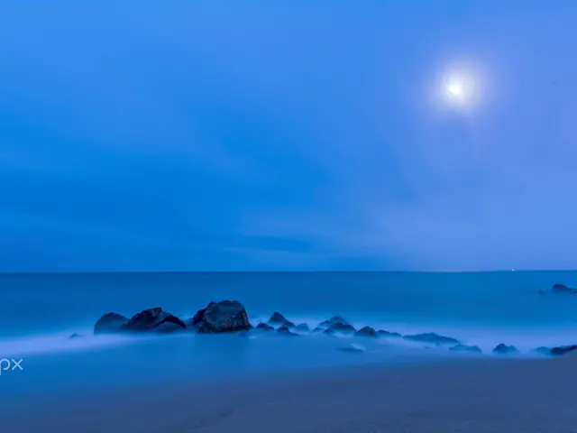 The image shows a long exposure shot of a full moon rising over the ocean, with rocks in the...