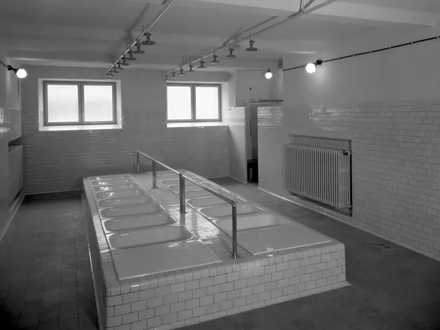 The image shows a black and white photo of a public restroom with a long row of sinks, taps, walls,...