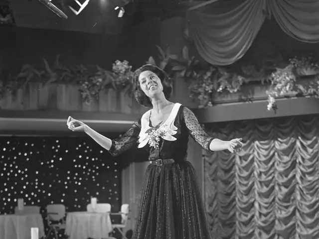 The image shows a woman in a black and white dress standing on a stage with a smile on her face. In...